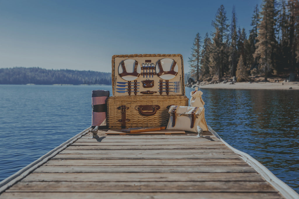 Wicker picnic basket with food items on a wooden dock by a lake with trees in the background