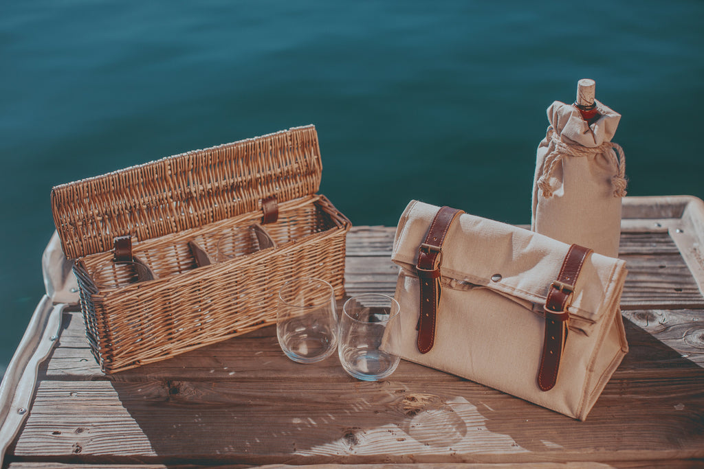 Wicker picnic basket, canvas bag, and wine bottle on a wooden dock by the water.
