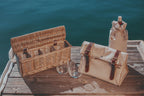 Wicker picnic basket, canvas bag, and wine bottle on a wooden dock by the water.