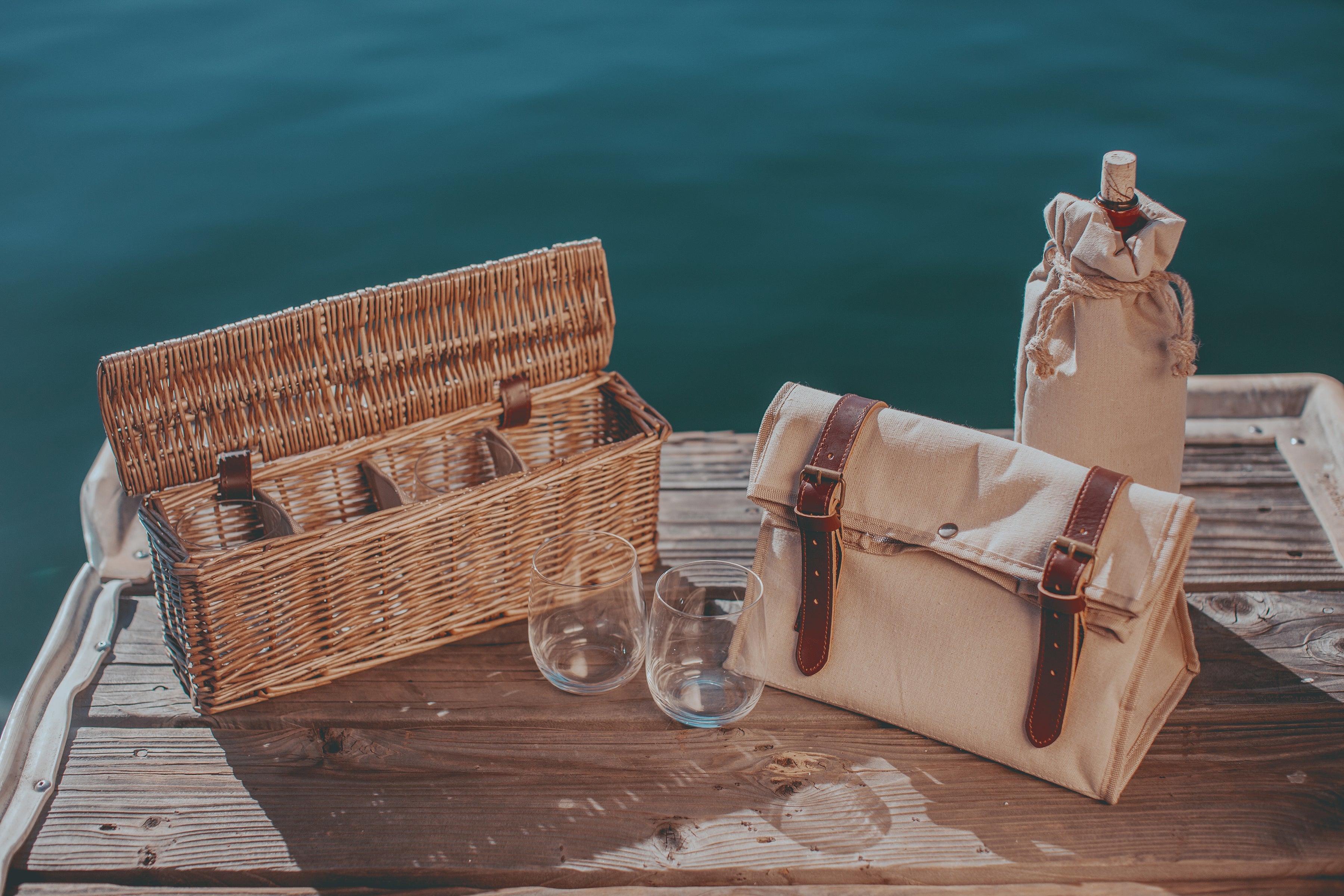Wicker picnic basket, canvas bag, and wine bottle on a wooden dock by the water.