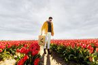 Person standing in a tulip field holding a cardboard box with a black arrow, wearing a yellow jacket and white pants.